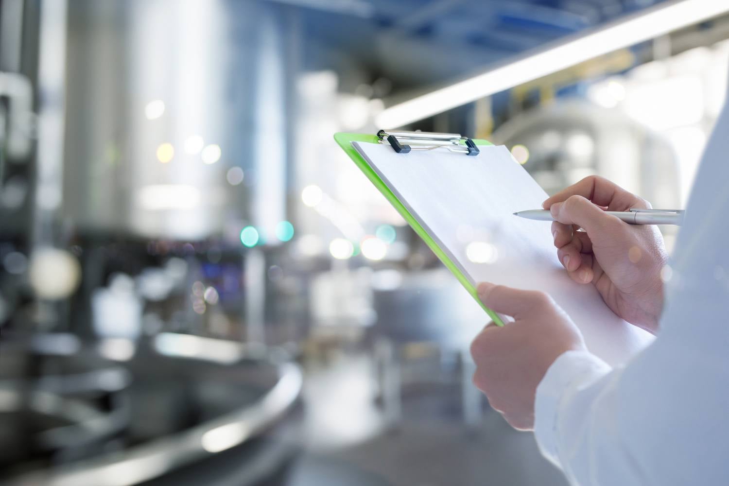 A person in a white lab coat holding a clipboard and pen in a factory setting.