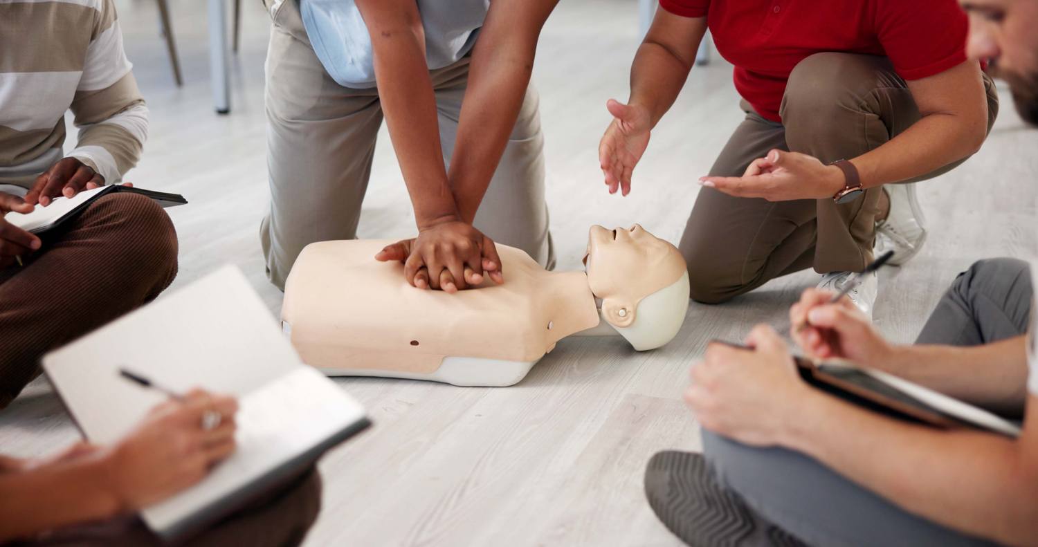 A group of people practising CPR on a mannequin with notepads in hand.