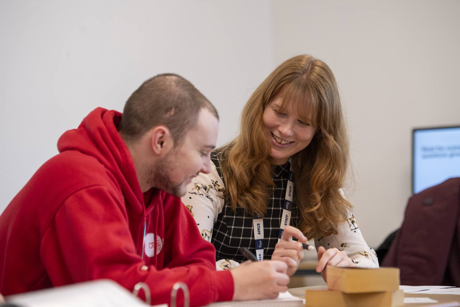 Two people in a classroom, one in a red hoodie and the other in a floral top, smiling and working together on a craft project with cardboard boxes.