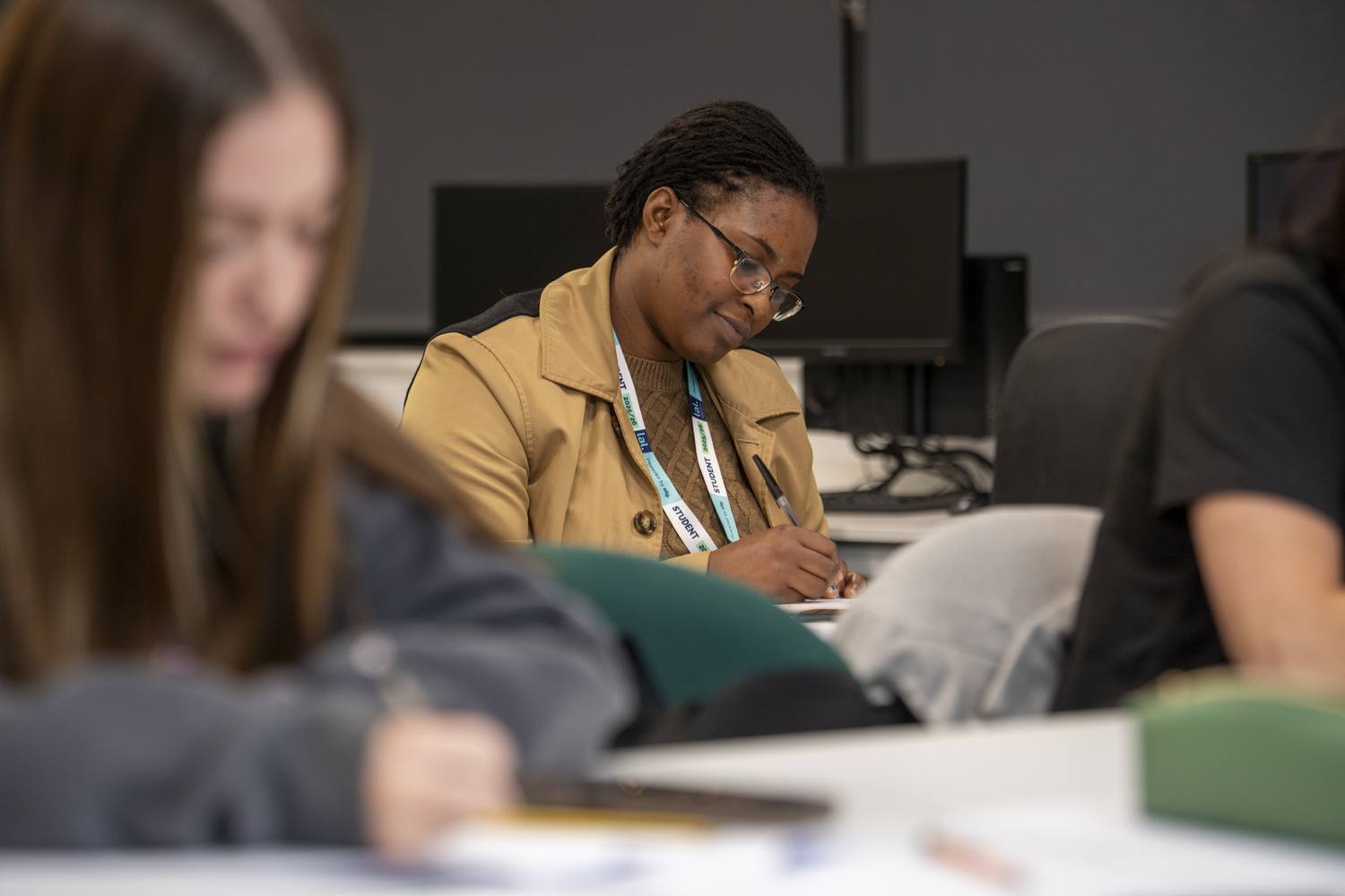 A woman in a tan jacket writing in a notebook at a table with other students in a computer lab.
