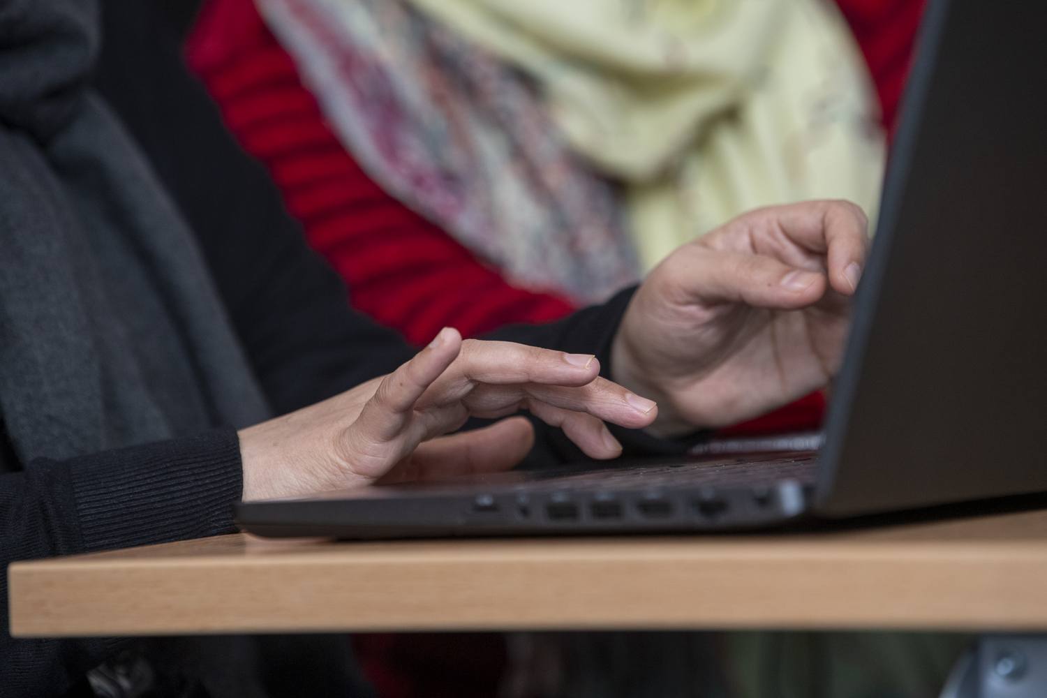 Two people using a laptop on a wooden table, focused on typing.