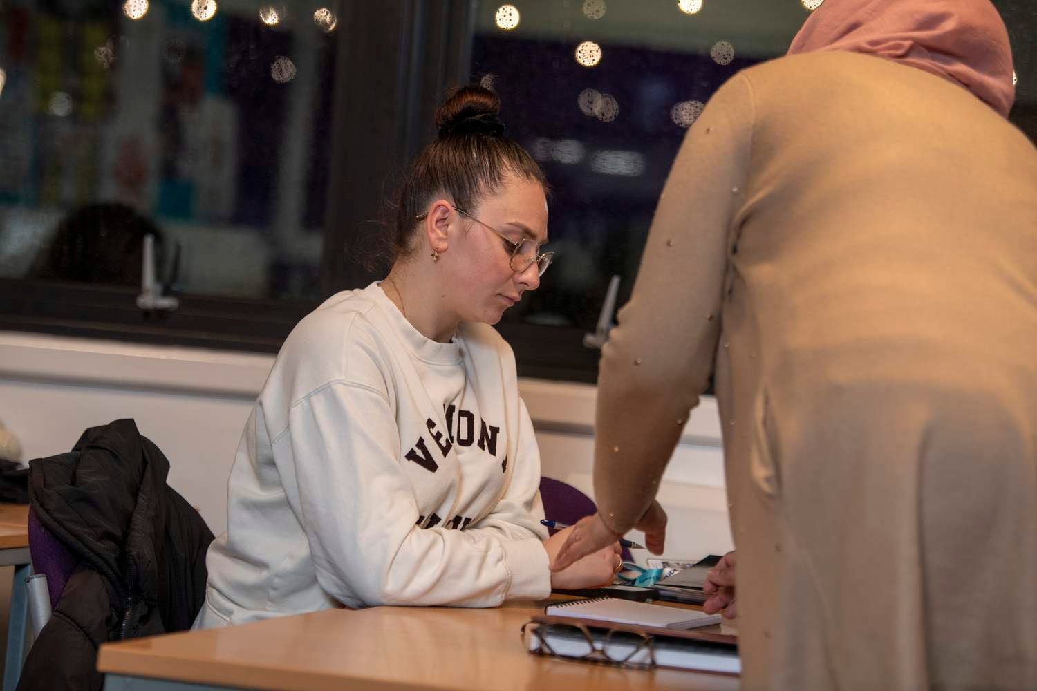 Two people at a desk, one writing in a notebook while the other points at something on the table.