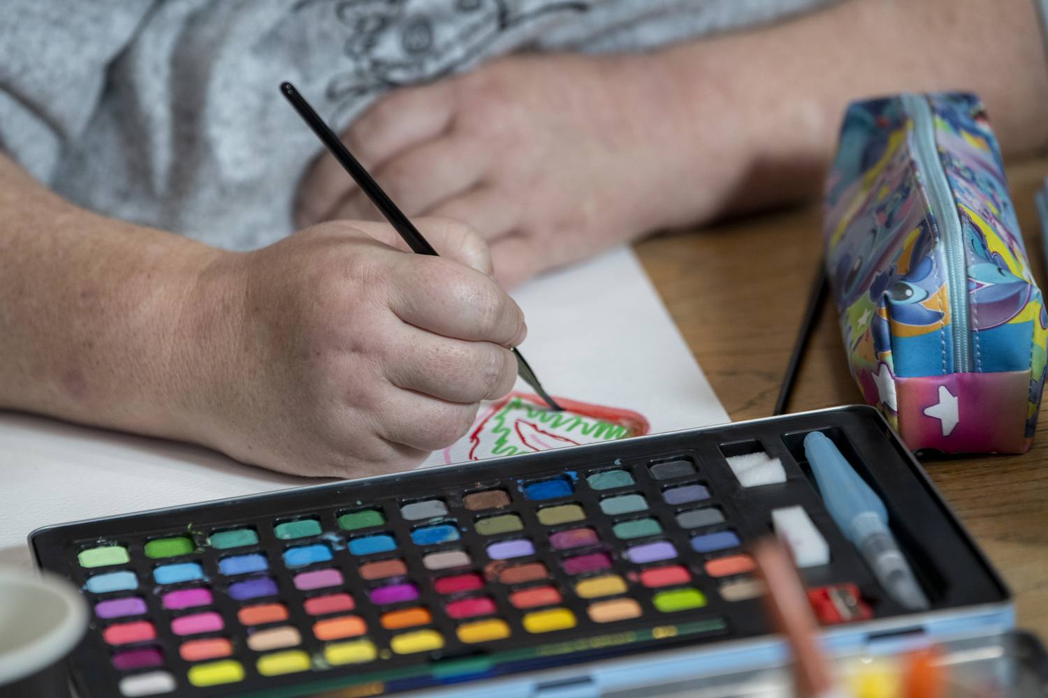 A person sketching with a black pen on paper, next to a colourful watercolor palette and a pencil case.