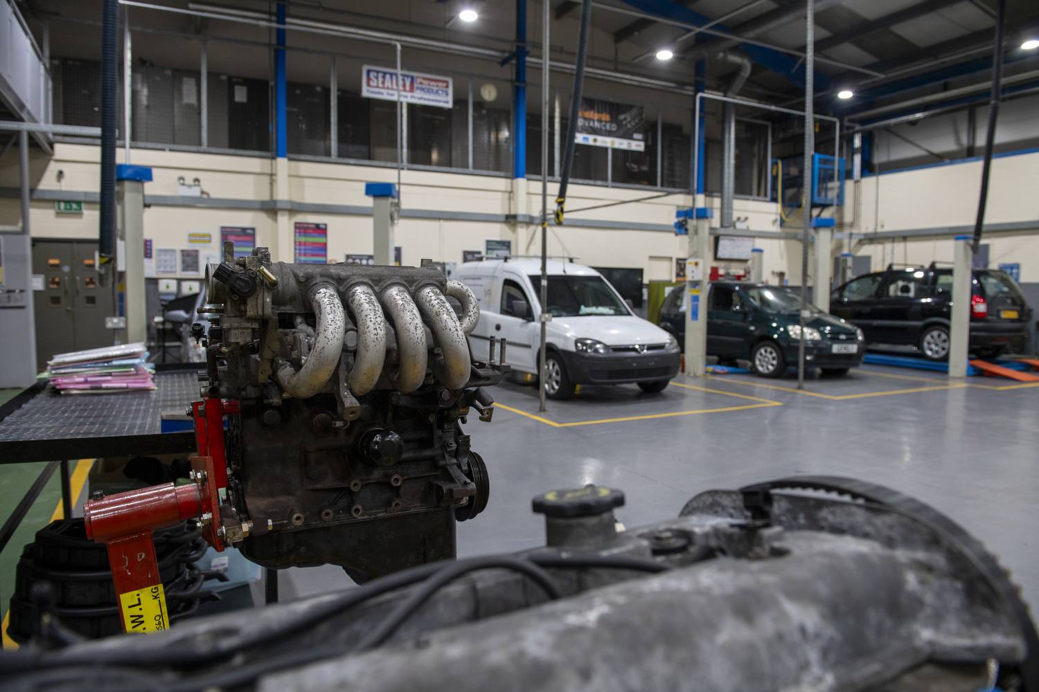 A V8 engine on a stand in a garage with cars and vans in the background.