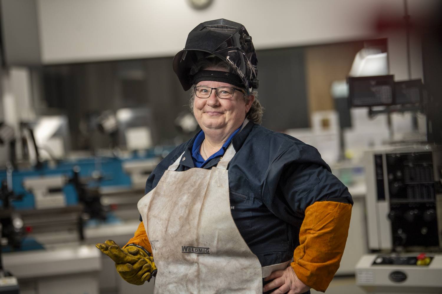 A woman in a welding helmet and apron stands confidently in a workshop, showcasing her skills in metalworking education.