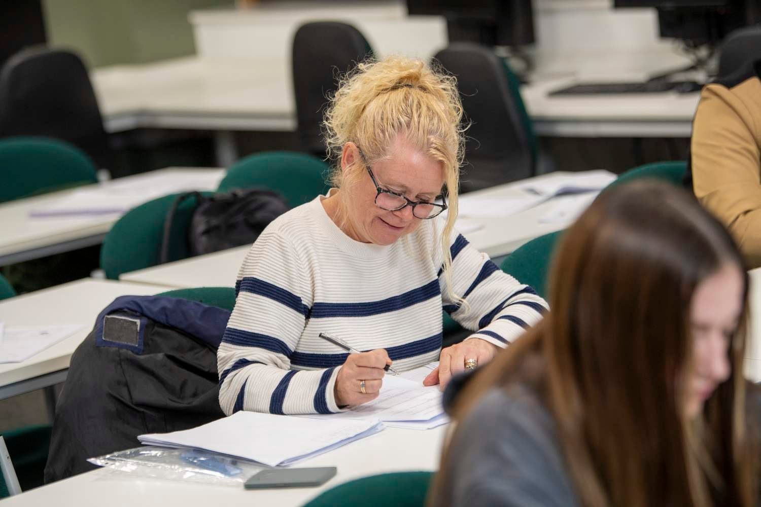 A woman in a striped jumper writing in a notebook at a table in a classroom.