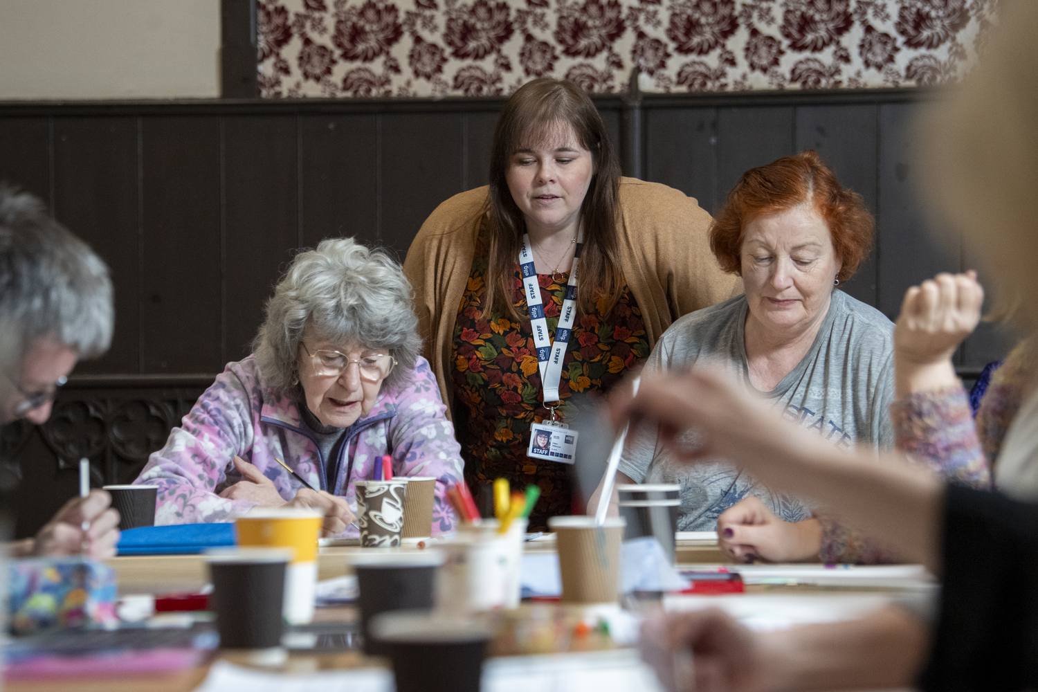 Three elderly women engaged in an art class, focused on drawing at a table with coloured pens and cups.