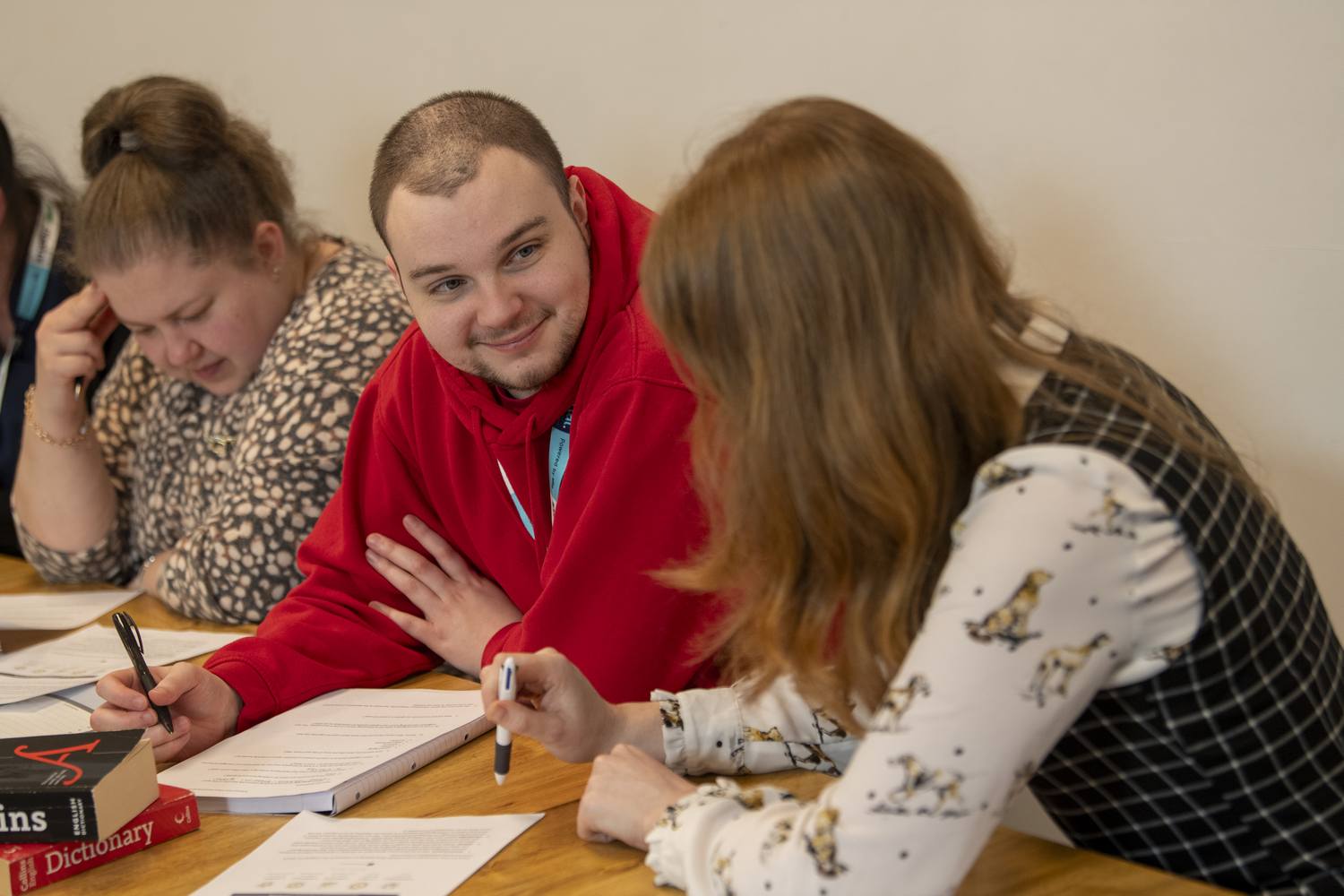 Three students in a classroom, engaged in a discussion. One student in a red hoodie writes in a notebook, while others listen attentively. A dictionary is visible on the table.