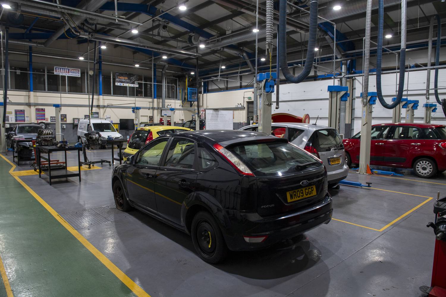 A college automotive workshop with several cars on lifts, tools, and equipment, under bright overhead lights.