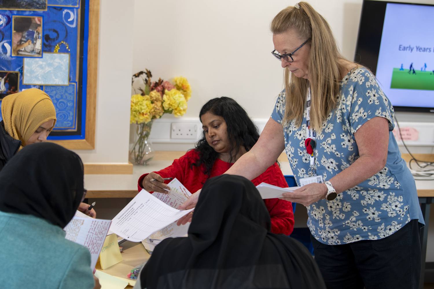 A woman in a floral dress assists seated students with paperwork in a classroom setting.