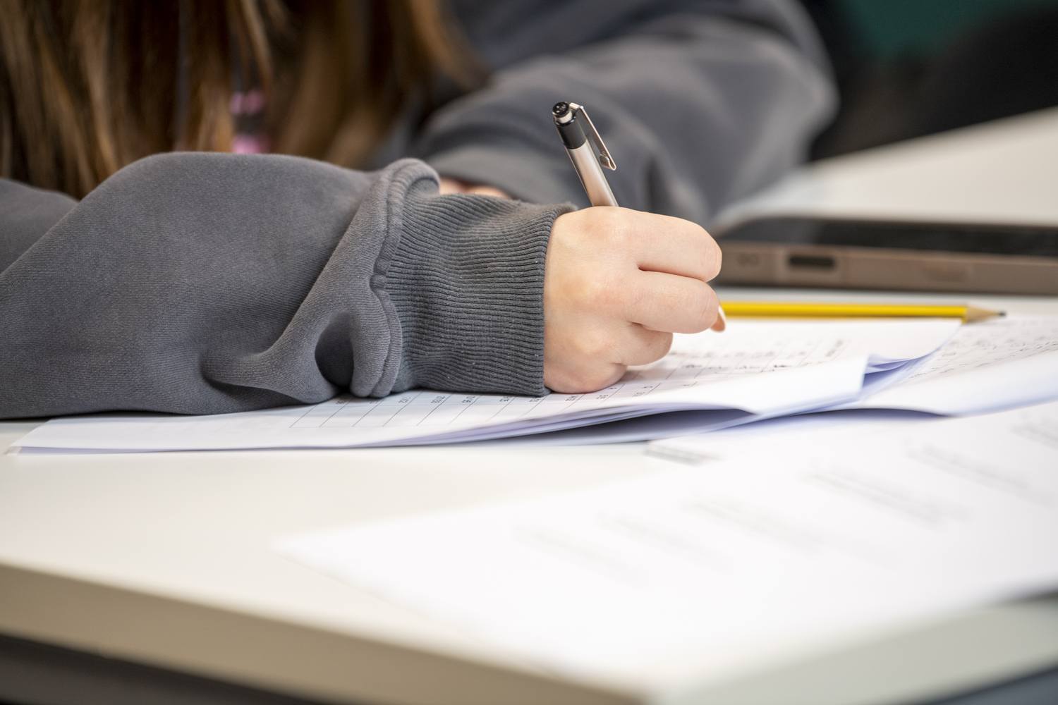 A student resting their head on a desk, holding a pen over an open notebook, with a laptop nearby.