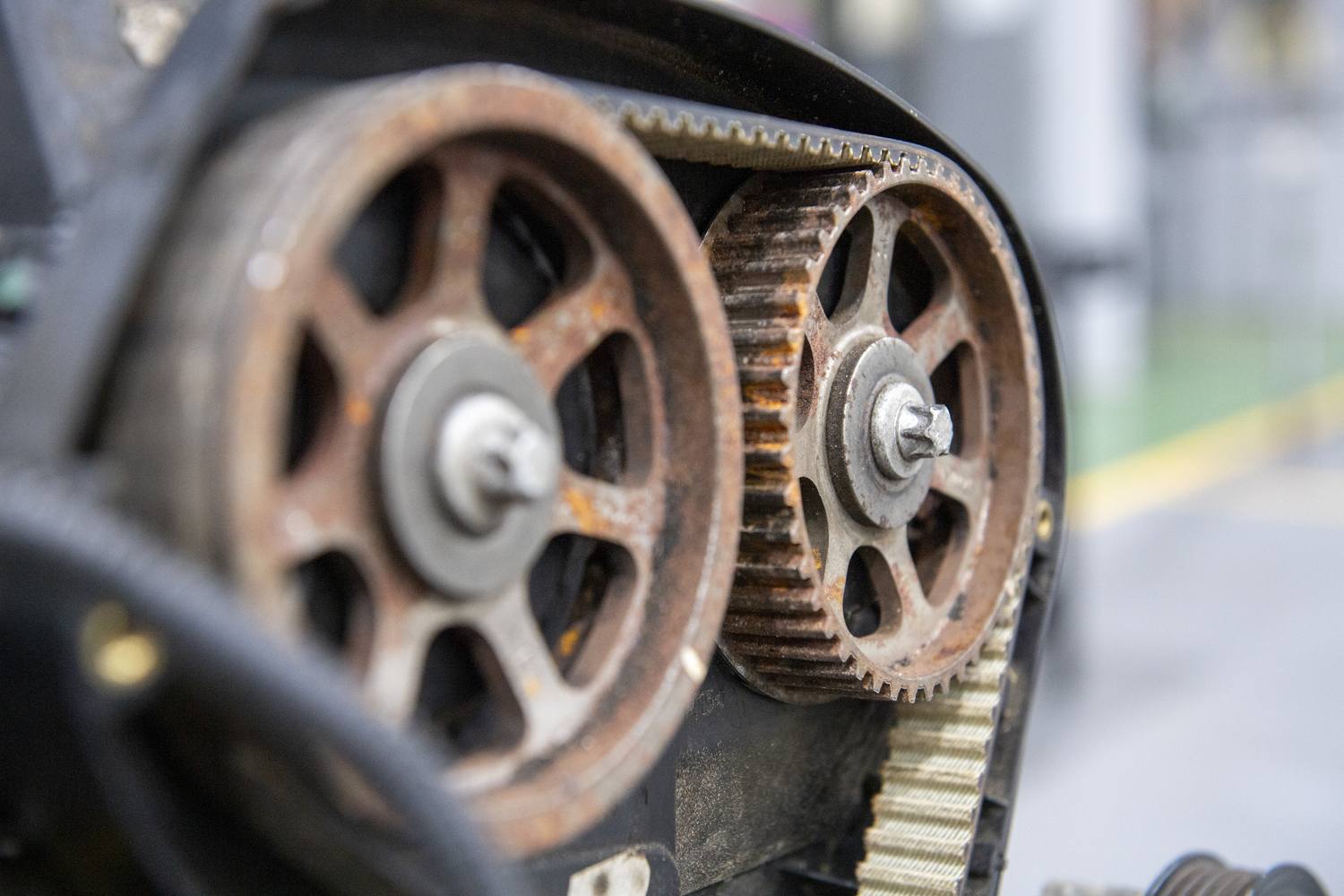 Two rusted metal gears with teeth and bolts, connected by a black belt, on a machine.