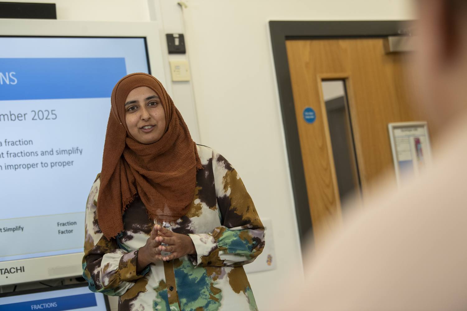 A woman in a patterned dress holding a glass of water stands in front of a projection screen displaying maths topics.