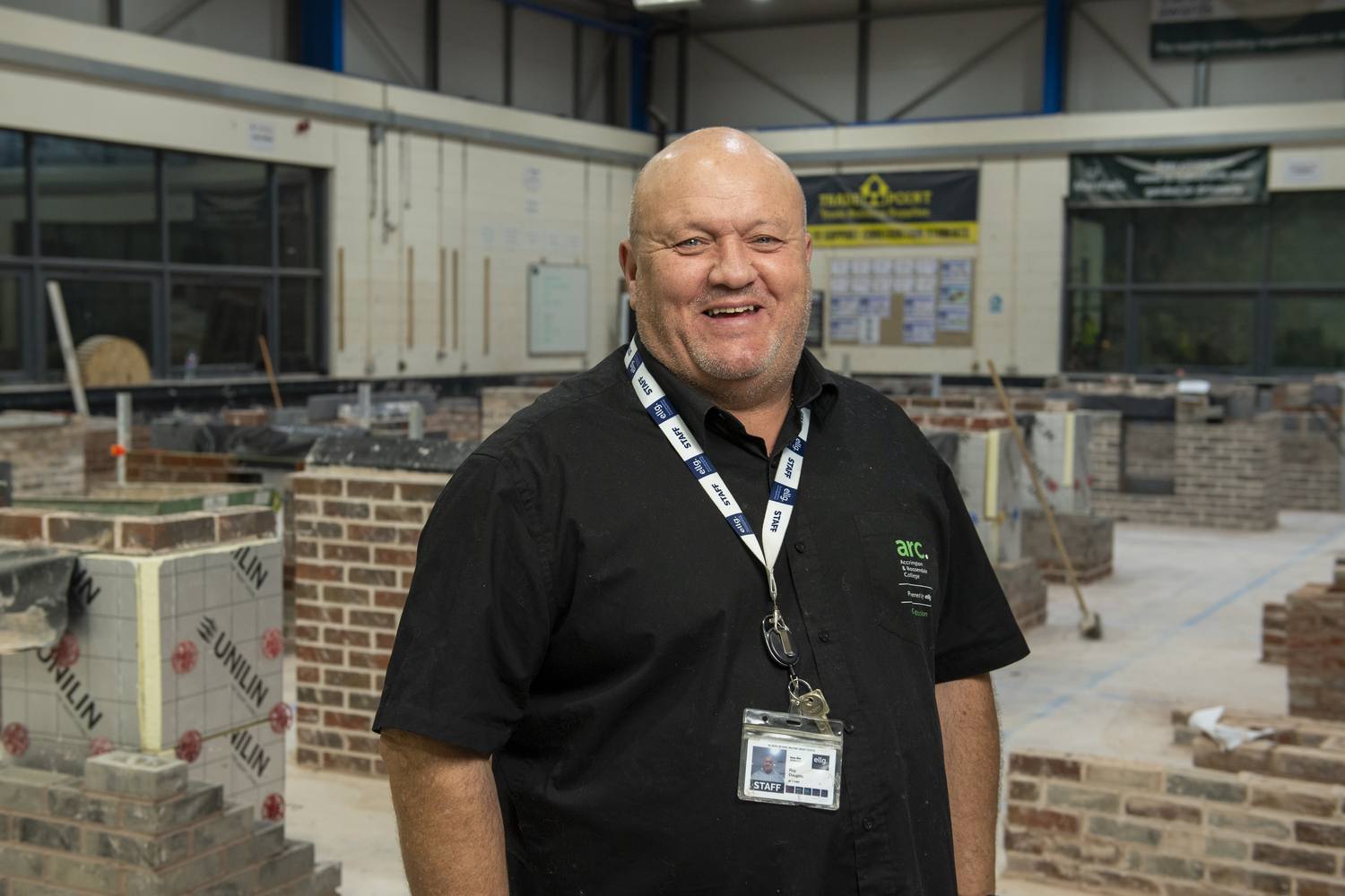 A man in a black polo shirt with a lanyard stands in front of brick walls in a college workshop.