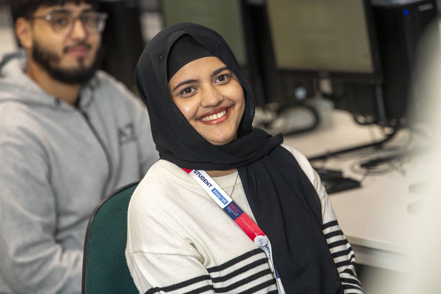 Two students smiling in a computer lab, wearing student lanyards, engaged in learning.