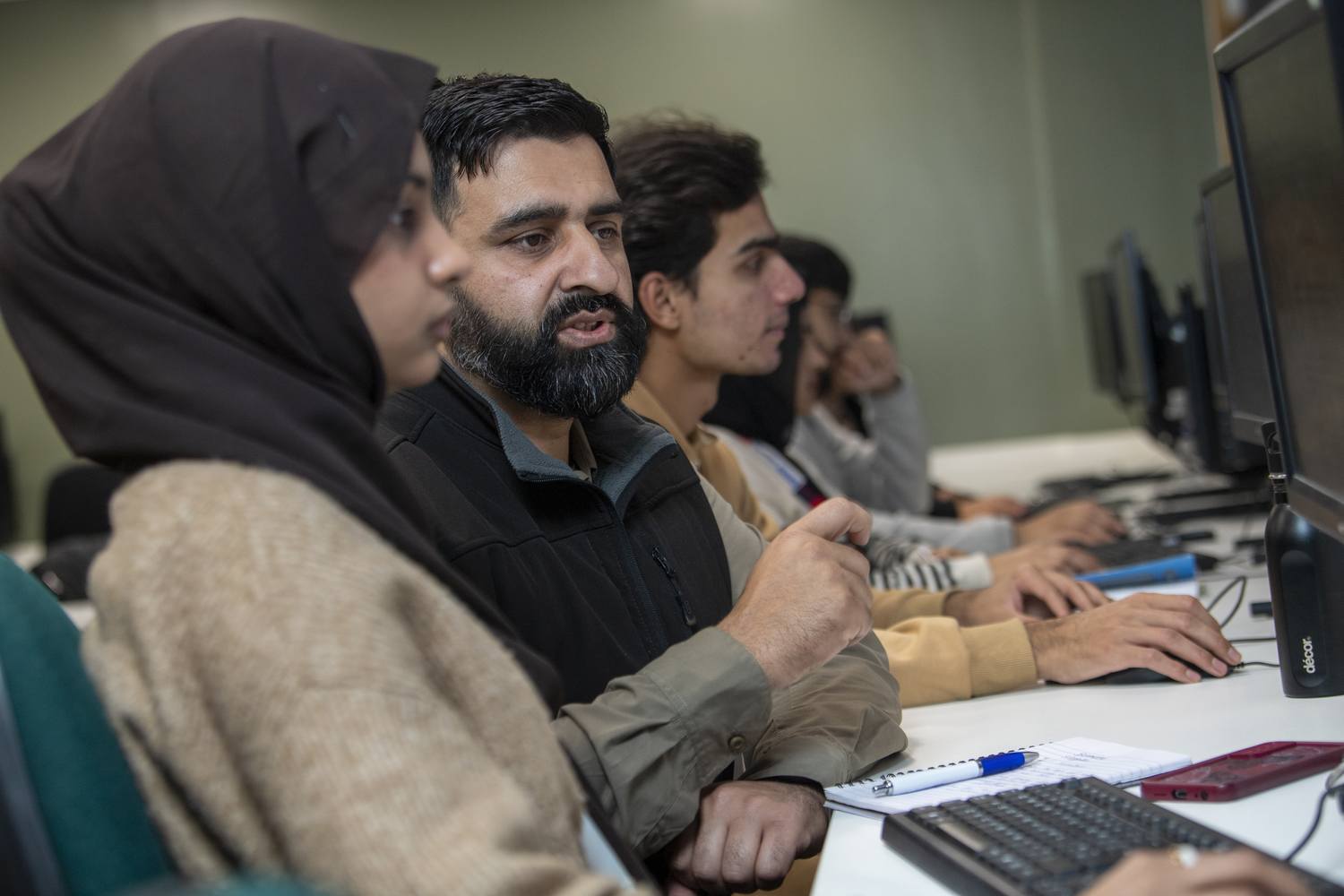 A group of students in a computer lab, focused on their screens, taking notes, and engaged in learning.