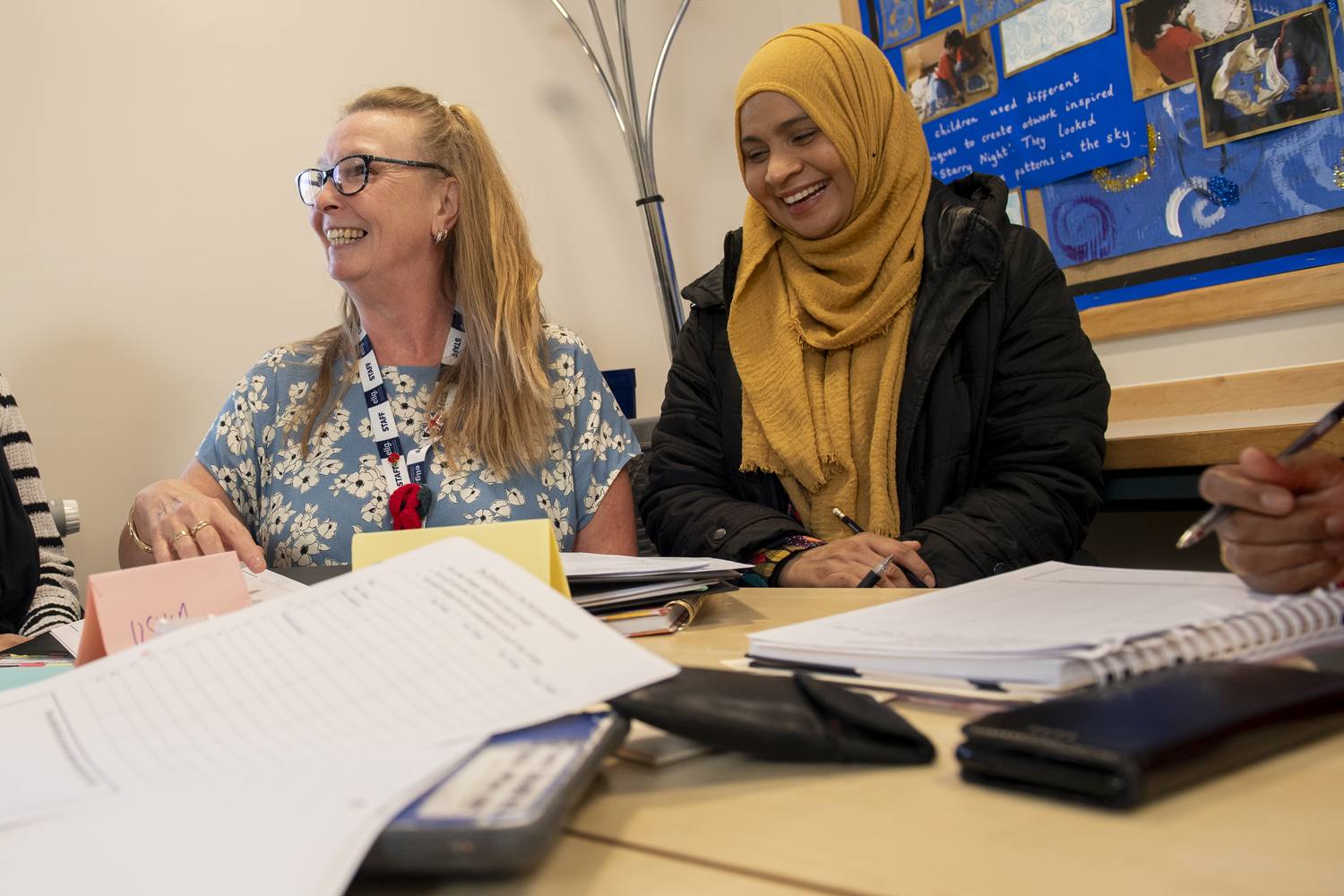 Two women smiling at a table with papers and a calculator, engaged in a discussion or workshop.