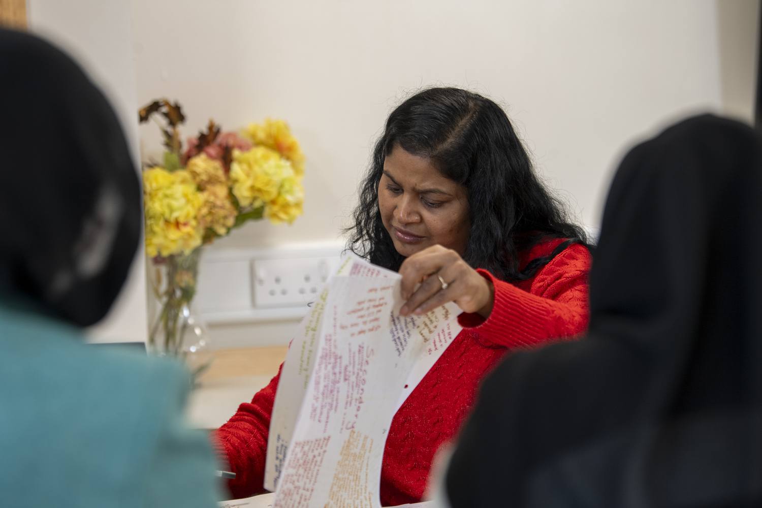 A woman in a red jumper reading a document at a table with flowers in the background.