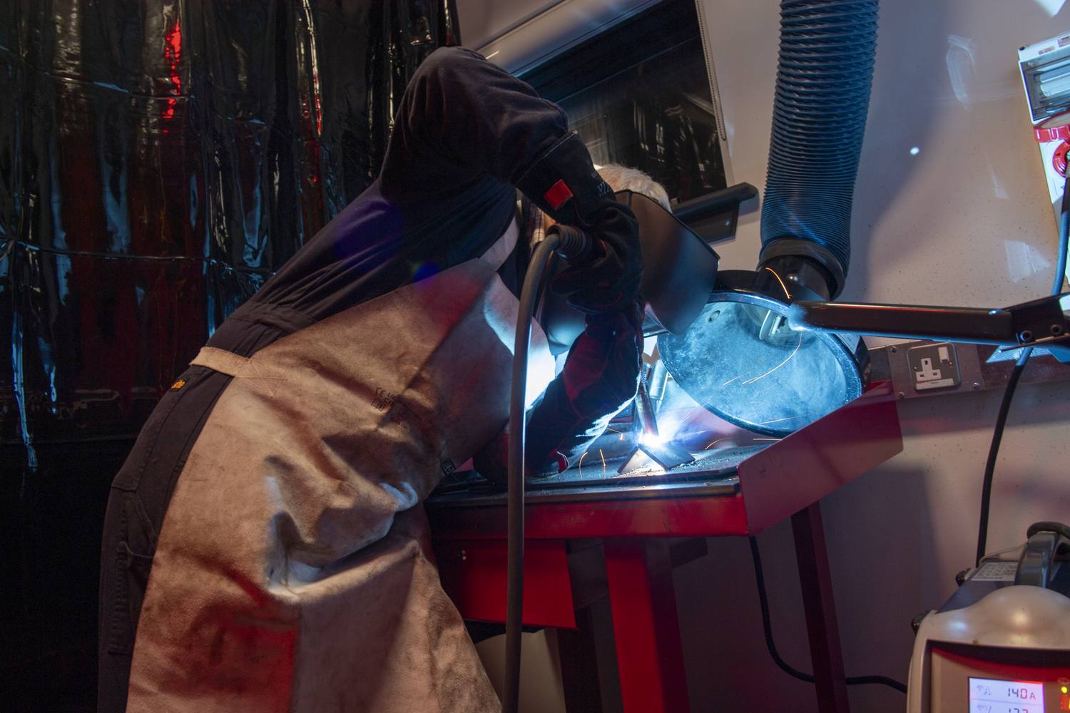 A person in protective gear welding a metal structure in a workshop, with bright blue sparks and a fume extractor nearby.