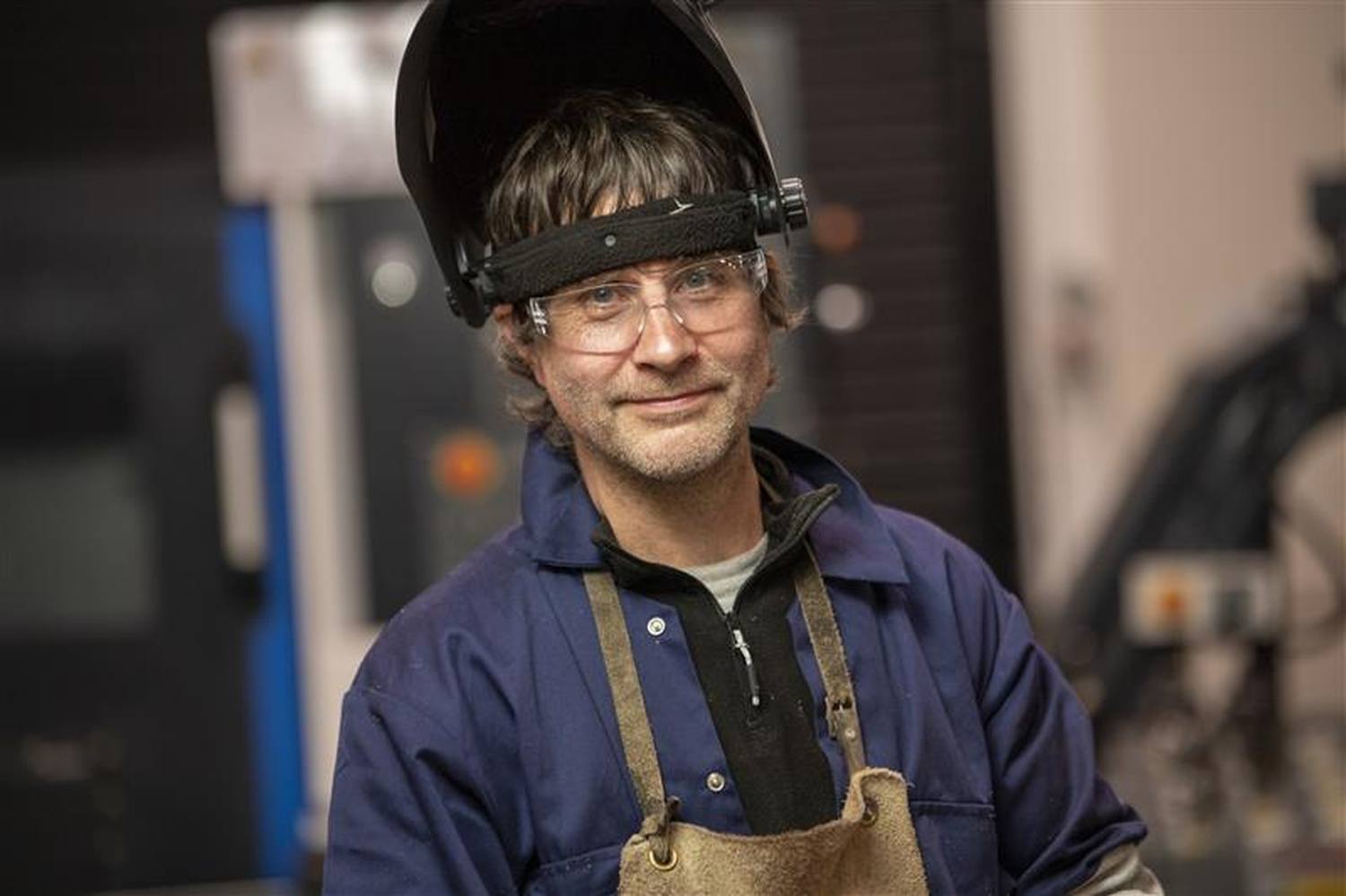 A man in a welding helmet and safety glasses smiling in a workshop, highlighting practical skills in engineering education.