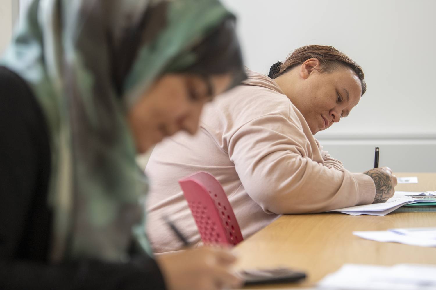 Two students in a classroom, focused on writing in notebooks at a wooden desk.