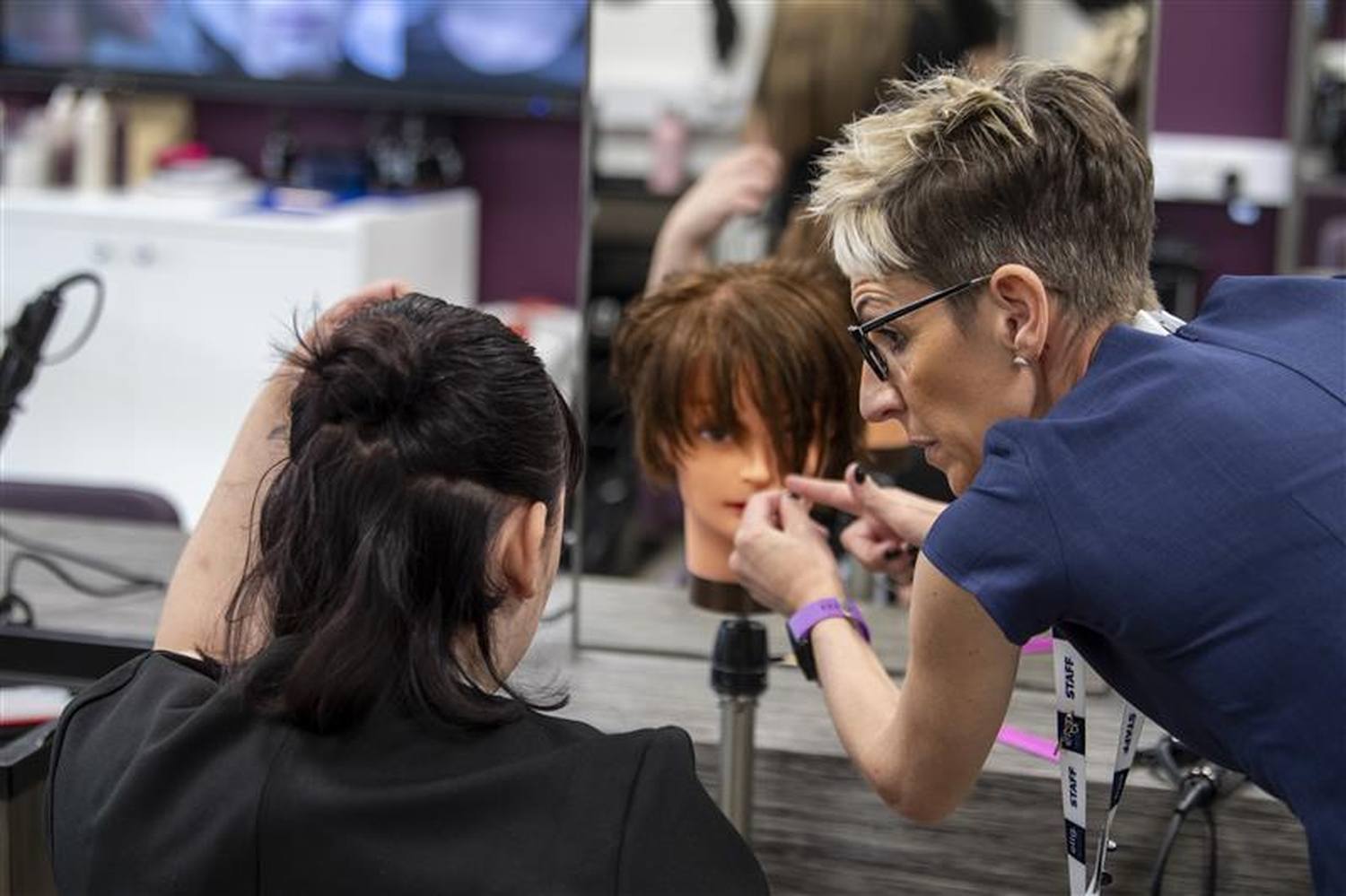 A woman in a blue shirt styles a mannequin's hair in a salon setting, demonstrating hairstyling techniques.