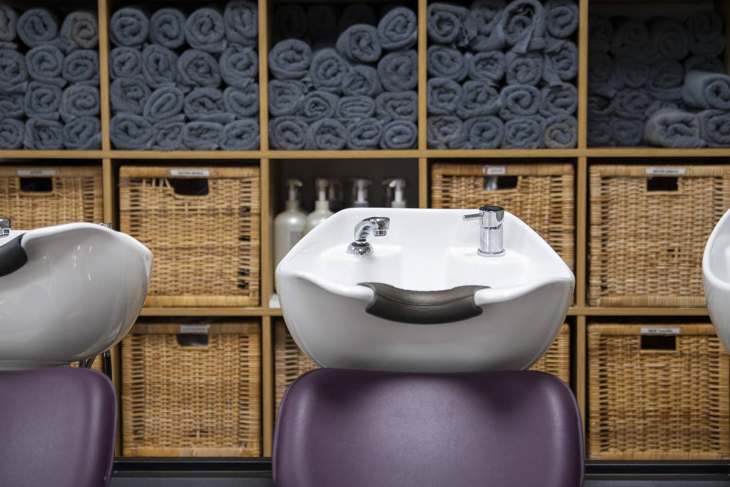 Three white hair washing sinks in front of wicker baskets with rolled-up grey towels.
