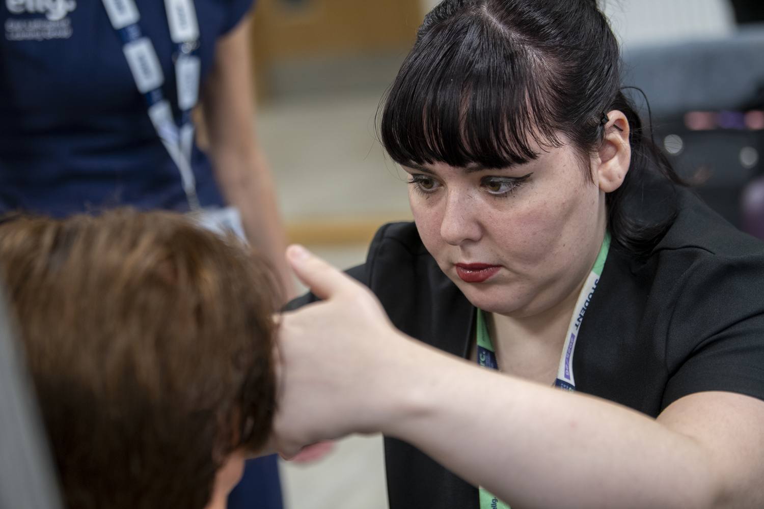 A woman in a black blazer examines a student's hair in a classroom setting, focusing on educational interaction.