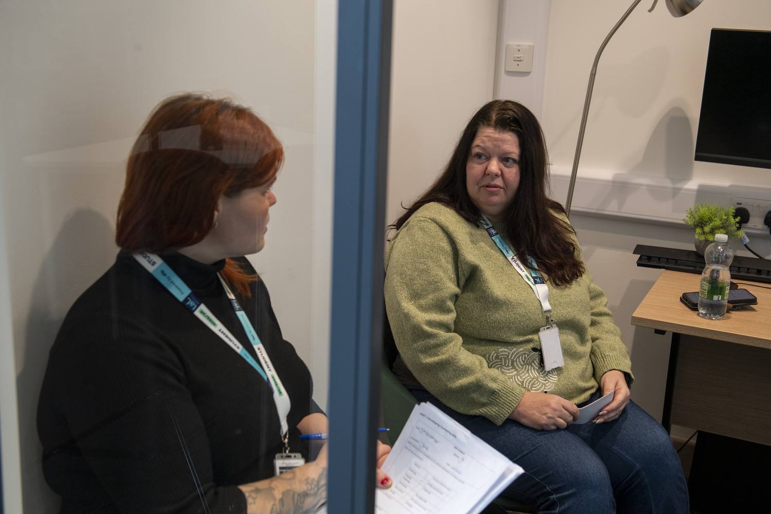 Two women in a consultation room, one wearing a face shield, discussing documents. A desk with a computer and water bottle is visible.