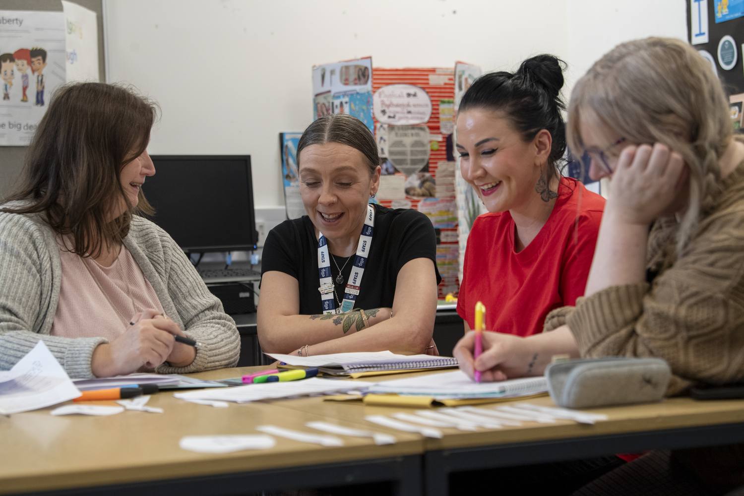 Four women smiling and working together at a table with notebooks, pens, and educational posters in a classroom setting.