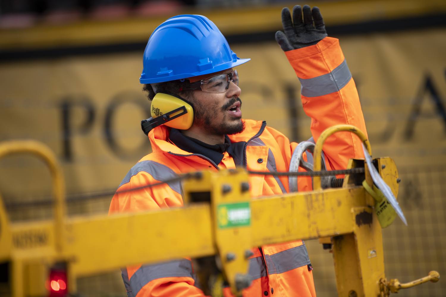 Construction worker in high-visibility gear and blue hard hat using a walkie-talkie on a yellow machine, gesturing with one hand.