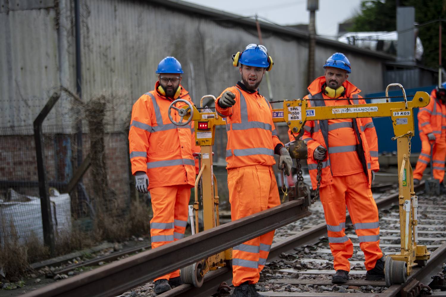 Three railway workers in high-visibility orange suits and blue hard hats operating manual track tools on railway tracks.