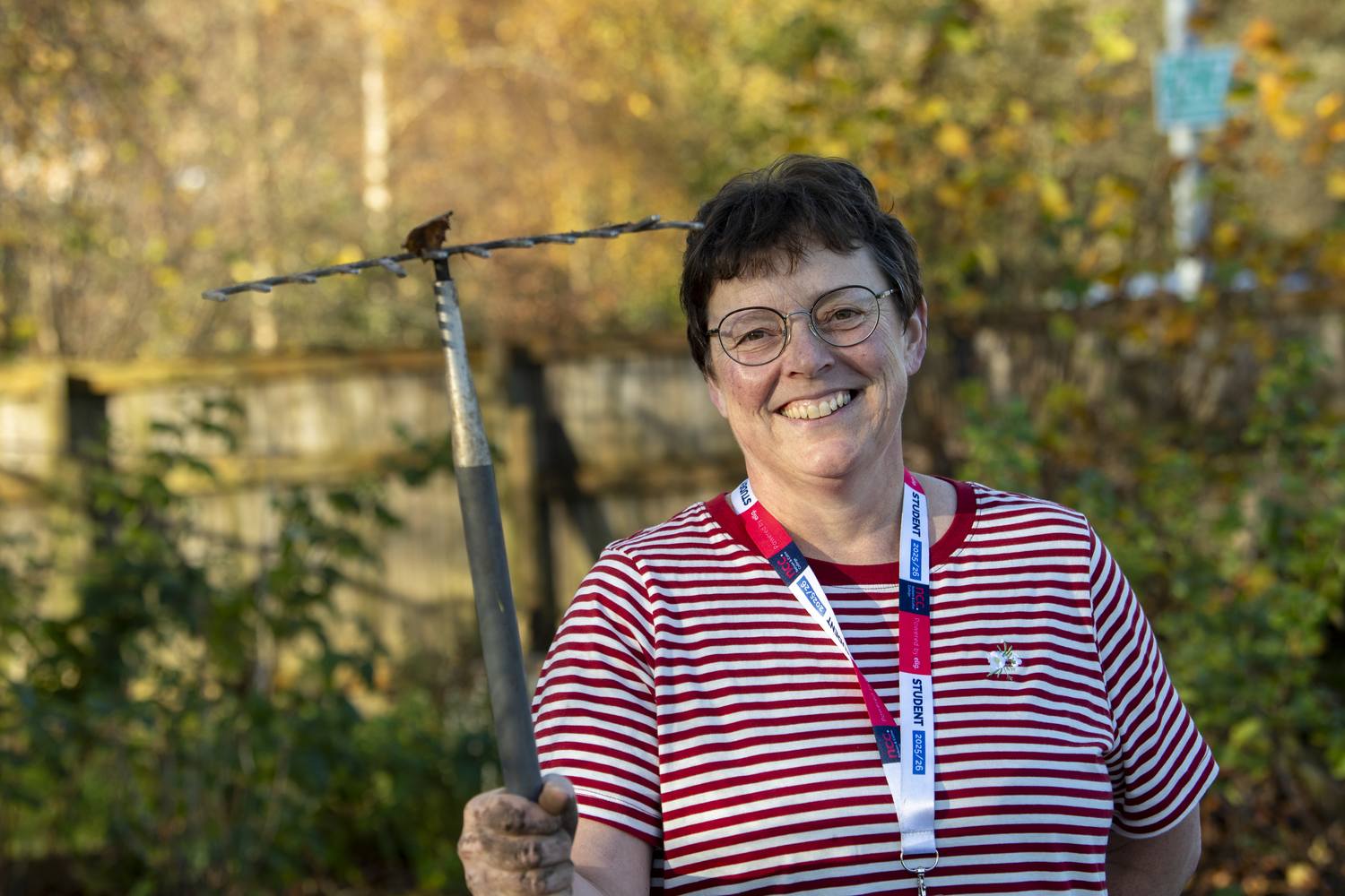 A woman in a red and white striped shirt holding a mattock in a garden, smiling at the camera.