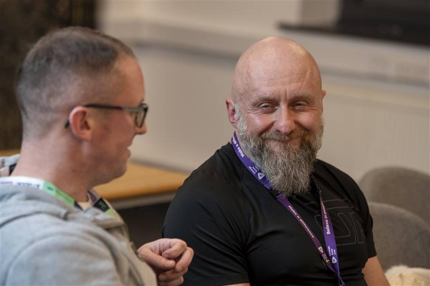 Two men in casual attire, one with glasses, engaged in conversation at a college event, wearing purple lanyards.