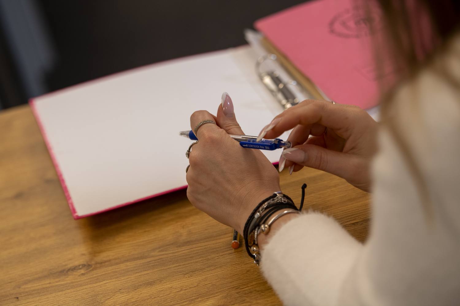 A person writing in a blue pen on a blank page in a pink binder on a wooden table.