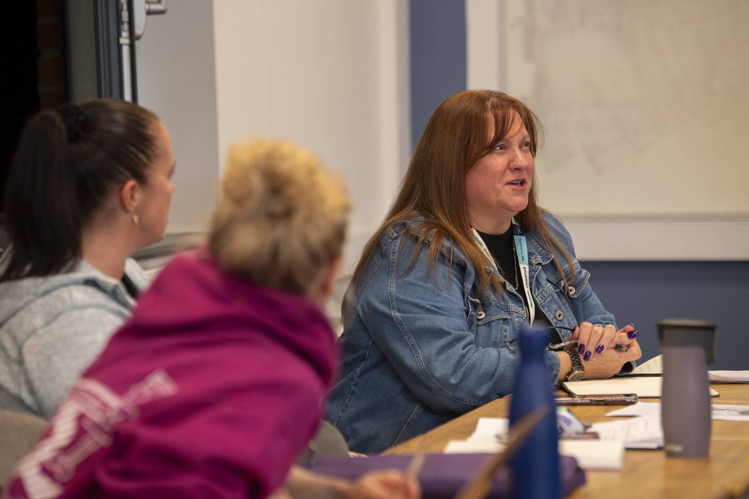 A woman in a denim jacket speaking at a table with three other women in a classroom setting.