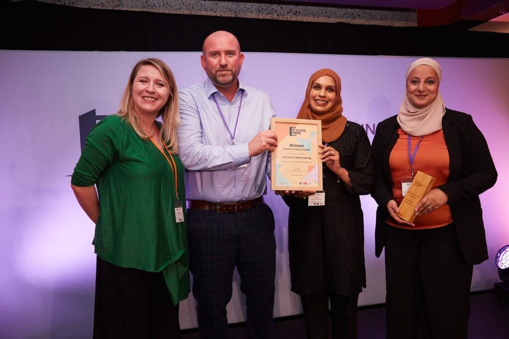 Four people on stage, two holding a certificate and one holding a wooden plaque, smiling at a college event.