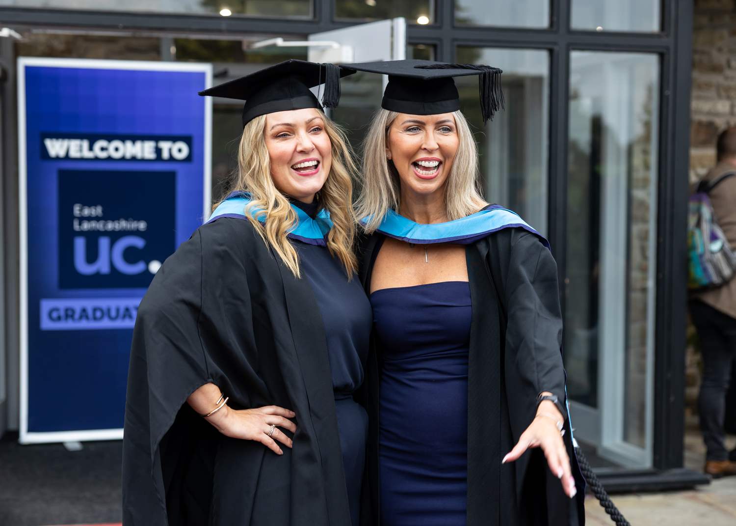 Two graduates in black gowns and caps smiling outside East Lancashire University Centre, celebrating their achievement.