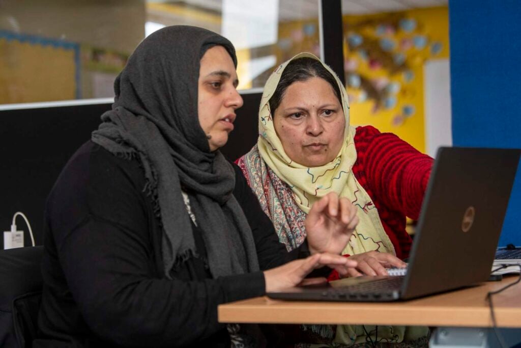 Two women in a classroom, one pointing at a laptop screen while the other watches attentively, engaged in a learning activity.