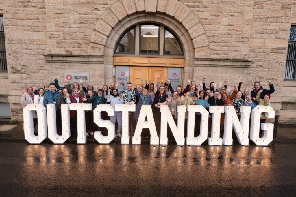 Group of people posing with large illuminated letters spelling 'OUTSTANDING' in front of a stone building.